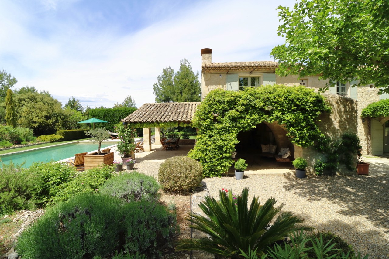 A serene backyard of a mediterranean-style home featuring a lush garden, an outdoor sitting area under an ivy-covered archway, and a swimming pool surrounded by trees.